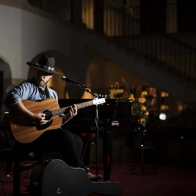 Mann mit Hut spielt Gitarre in dunklem Raum bei Mikrofon