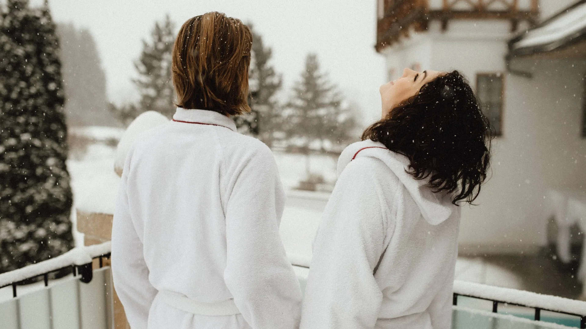 Zwei Personen in weißen Bademänteln genießen Schneefall auf einem Balkon